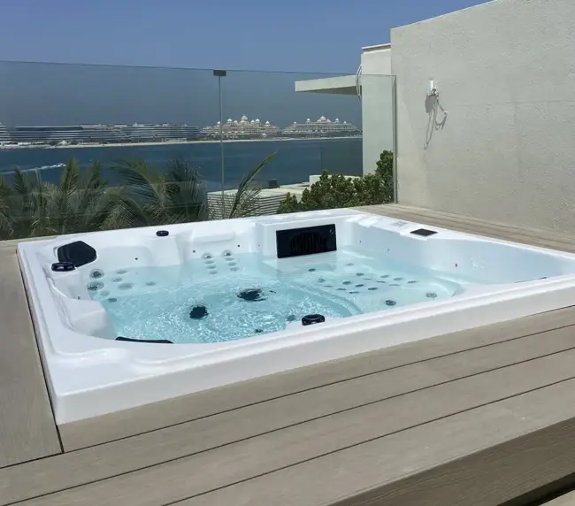 A modern jacuzzi on a wooden decking overlooks the sea in Dubai, with palm trees, cruise ships, and a clear blue sky in the background. A glass balustrade lines the edge of the decking.