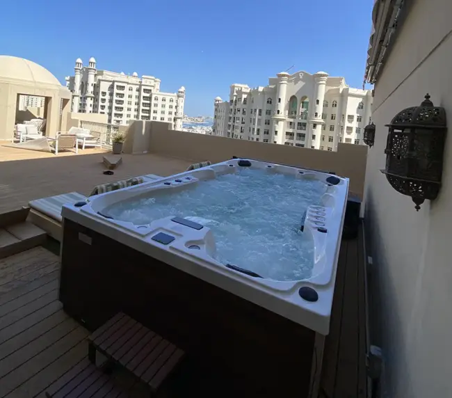 A bubbling jacuzzi on a rooftop terrace, surrounded by loungers. Ornate buildings with arched windows and balconies are visible in the background under a clear blue sky.