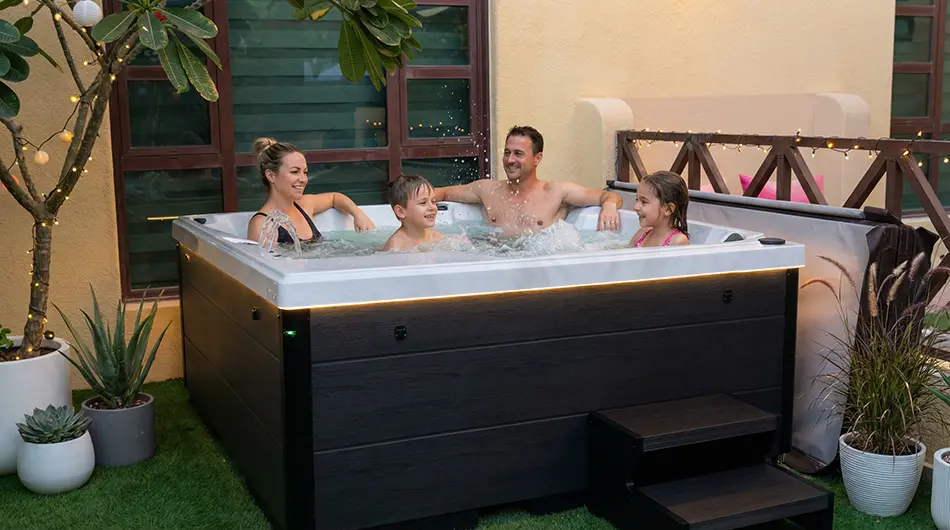 Four people, two adults and two children, relax and smile together in a hot tub jacuzzi surrounded by plants and fairy lights in a cosy outdoor patio setting.