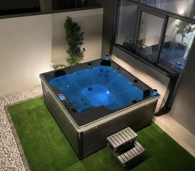 A square jacuzzi with glowing blue water sits on artificial grass in a modern Abu Dhabi outdoor patio area, next to a window, with a small step and decorative white stones on one side.