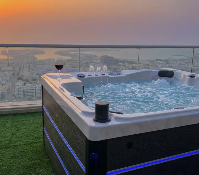 A bubbling jacuzzi on a rooftop terrace overlooks the city and sea at sunset, with a glass of red wine on the tub’s edge and artificial grass underfoot—perfect for relaxing in this luxurious flat rental.