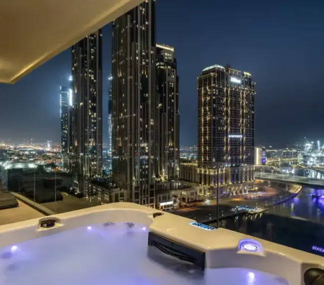 A rooftop jacuzzi at night on a balcony overlooks Dubai's illuminated skyscrapers and the river below, creating a luxurious urban atmosphere.